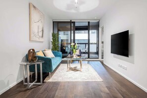 Una sala moderna con un sofá azul, una mesa de centro de cristal, un televisor de pantalla plana y un ventanal corredero que da a un balcón.