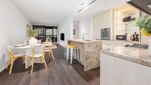 A modern kitchen with a marble island, a dining area with white chairs, and a large window. - LaneStay - 2BR Refined Living, Pool & Gym & Parking (Melbourne)