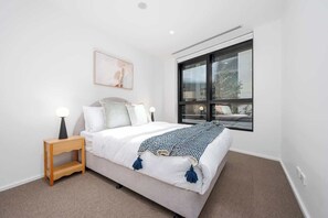 Een slaapkamer met een bed, nachtkastje, lamp, raam en een kunstwerk.