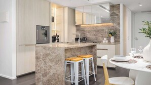 A modern kitchen with a marble island, white cabinetry, and a dining area with a white table and chairs. - LaneStay - 2BR Refined Living, Pool & Gym & Parking (Melbourne)