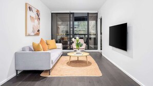 A modern living room with a grey sofa, a coffee table, a flat-screen TV, and a large window. - LaneStay - 2BR Refined Living, Pool & Gym & Parking (Melbourne)