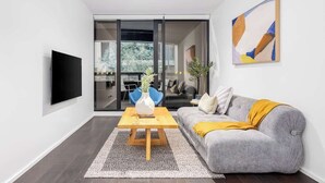 A modern living room with a grey sofa, a wooden coffee table, a flat-screen TV, and a sliding glass door leading to an outdoor area. - LaneStay - Grand Prix Retreat w Pool, Gym, Parking (Melbourne)