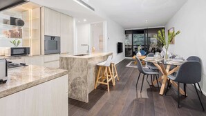 A modern kitchen with a central island, dining area with chairs, and a large window. - LaneStay - Grand Prix Retreat w Pool, Gym, Parking (Melbourne)