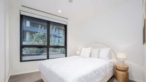 A bedroom with a large window, a bed, a nightstand, and a lamp. - LaneStay - Grand Prix Retreat w Pool, Gym, Parking (Melbourne)