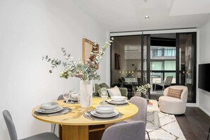 A dining area with a round wooden table set for four, a vase with greenery, and a modern interior with a sofa and a television. - LaneStay - Contemporary Living, Pool, Gym, Parking (Melbourne)