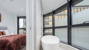 Ein modernes Badezimmer mit einer großen, verglasten Duschkabine, einer weißen Badewanne und einem Bett mit braunem Kopfteil.