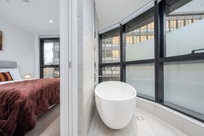 Ein modernes Badezimmer mit einer großen, verglasten Duschkabine, einer weißen Badewanne und einem Bett mit braunem Kopfteil.
