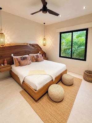 A modern bedroom with a wooden headboard, a large bed, two round woven ottomans, a woven rug, and a window with a view of greenery. - Casa Entre Monos (Hacienda Pinilla)