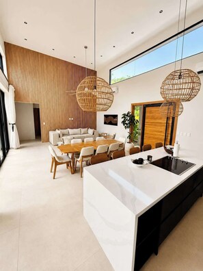 A modern kitchen with a central island, a dining area with a wooden table and chairs, and a living space with a sofa and a large window. - Casa Entre Monos (Hacienda Pinilla)