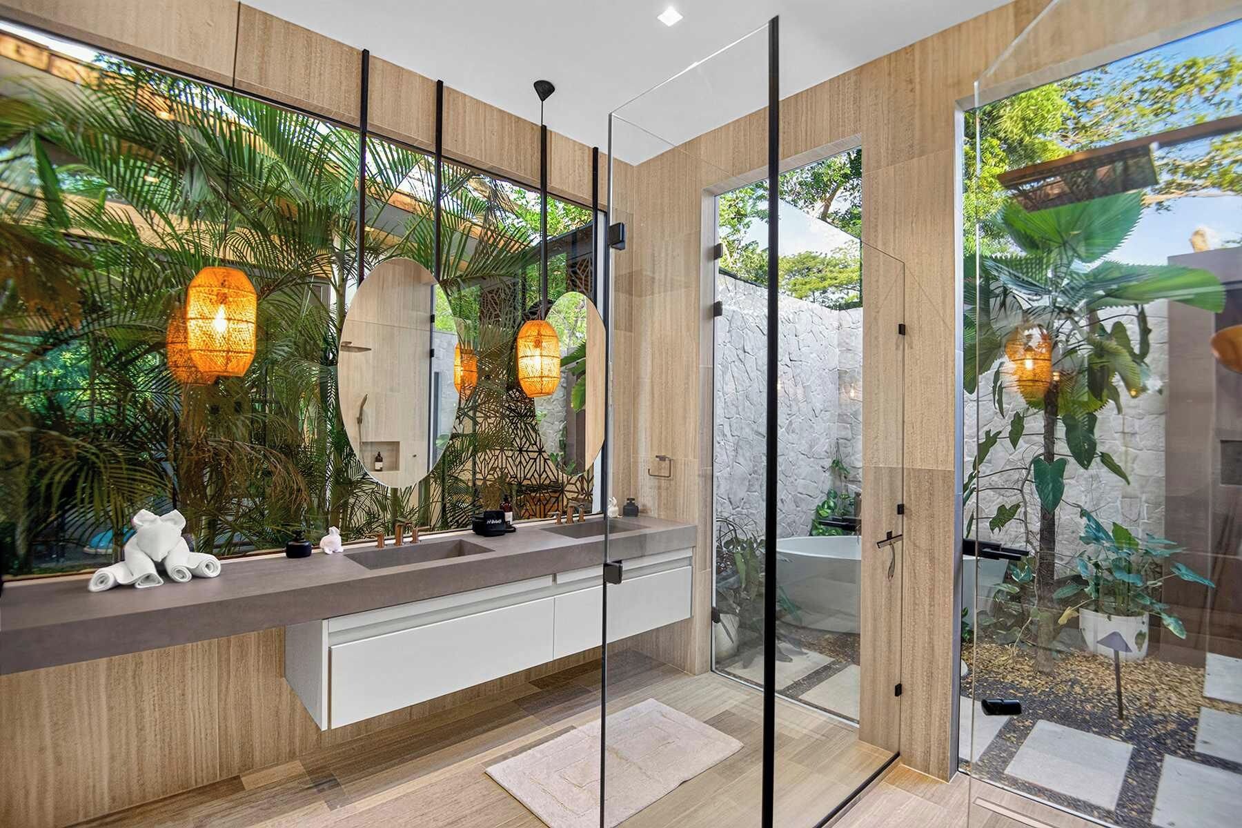 A modern bathroom with a wooden vanity, large mirrors, and a view of a tropical garden.