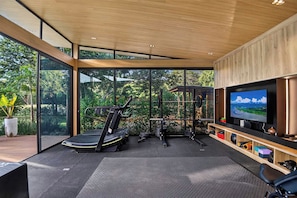 A modern home gym with exercise equipment, a television, and large windows offering a view of the outdoors. - Casa Kaikoo (Hacienda Pinilla)