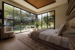 Ein Schlafzimmer mit einem großen Bett, einem Sofa und Blick auf einen Garten durch raumhohe Fenster.