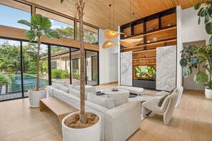 Ein modernes Wohnzimmer mit einem großen Ecksofa, einem Couchtisch und einem Fernseher. Ein großes Fenster bietet Blick auf einen Pool und viel Grün draußen.