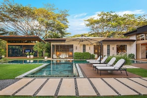 A modern outdoor pool area with lounge chairs, a stone deck, and a house with large windows and a stone facade. - Casa Kaikoo (Hacienda Pinilla)