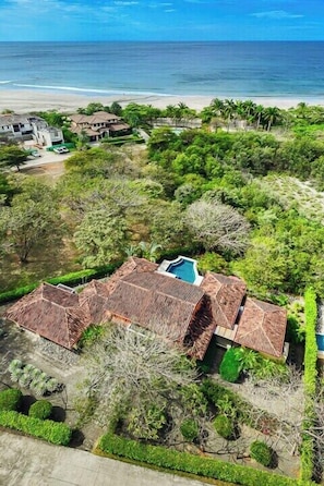An aerial view of a property with a swimming pool, surrounded by lush greenery and a beach.