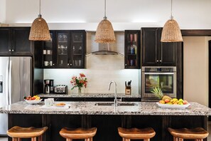 Eine Küche mit dunklen Schränken, einer Arbeitsplatte aus Granit, einem Edelstahl-Kühlschrank, einem Toaster, einer Spüle und drei hängenden Korbtropfenleuchten.