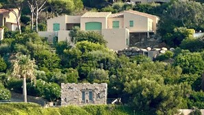 En fastighet vid kusten med arkitektur i medelhavsstil, inklusive en byggnad i sten och moderna hus med gröna fönsterluckor, omgiven av frodig grönska och berg i bakgrunden.