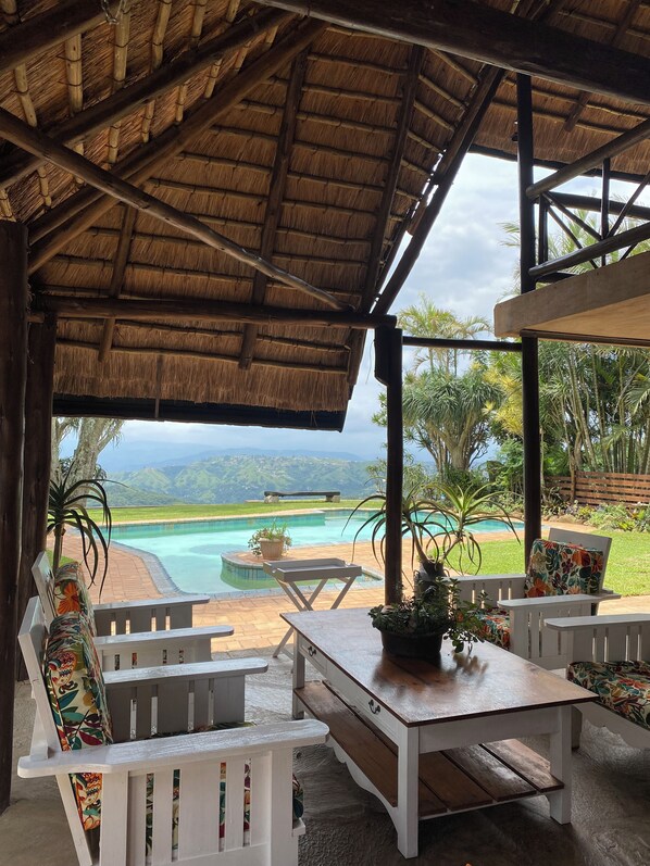 An outdoor seating area with a thatched roof, white furniture, and a view of a swimming pool and landscape.