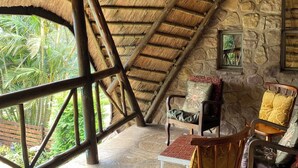 A room with a thatched roof, stone walls, a wooden chair, a table, and a window with a view of greenery. - Kariki Villa with Panoramic Valley Views (Outer West Durban)