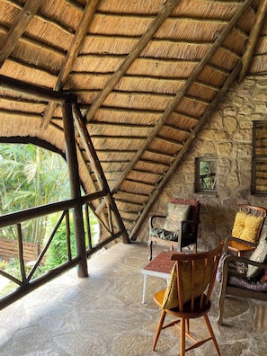 A room with a thatched roof, stone walls, a wooden chair, a table, and a window with a view of greenery.
