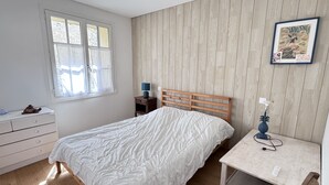 Un dormitorio con una cama de madera, una mesita de noche con lámpara, un aparador y una ventana con cortinas.