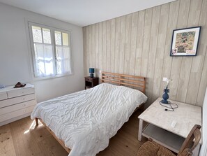 Ein Schlafzimmer mit einem hölzernen Bett, einem Nachttisch mit Lampe, einer Kommode und einem Fenster mit Vorhängen.