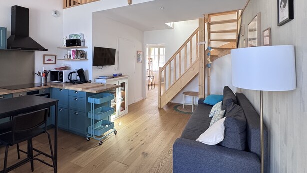 Una cocina moderna con microondas, tostadora y armarios, un comedor con mesa y sillas, una sala de estar con sofá y lámpara, y una escalera de madera.