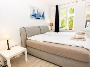 Ein Schlafzimmer mit Bett, Nachttisch, Lampe, einem Fenster mit Vorhängen und einem Bild an der Wand.