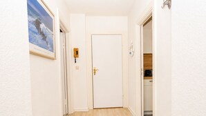 Ein Flur mit einer weißen Tür, einem gerahmten Bild an der Wand, einem Lichtschalter und einem offenen Durchgang zu einer Küche.