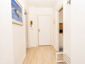 Ein Flur mit einer weißen Tür, einem gerahmten Bild an der Wand, einem Lichtschalter und einem offenen Durchgang zu einer Küche.