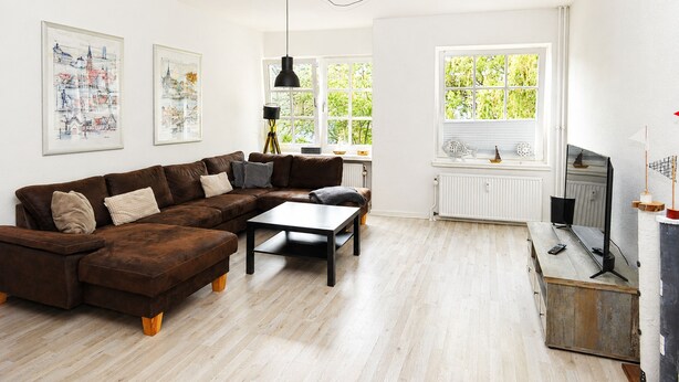 Ein modernes Wohnzimmer mit einem braunen Ecksofa, einem Couchtisch, einem Fernseher auf einem Holzboden und einem großen Fenster mit Blick auf Grünflächen.