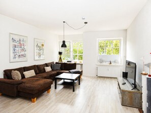 Ein modernes Wohnzimmer mit einem braunen Ecksofa, einem Couchtisch, einem Fernseher auf einem Holzboden und einem großen Fenster mit Blick auf Grünflächen.