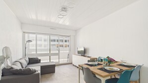 En moderne stue med sofa, spisebord med stole, fjernsyn og et stort vindue.