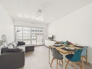Ein modernes Wohnzimmer mit einer Couch, einem Esstisch mit Stühlen, einem Fernseher und einem großen Fenster.