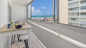 En balkon med udsigt over stranden og havet, et bord med stole og et vindue med persienner.