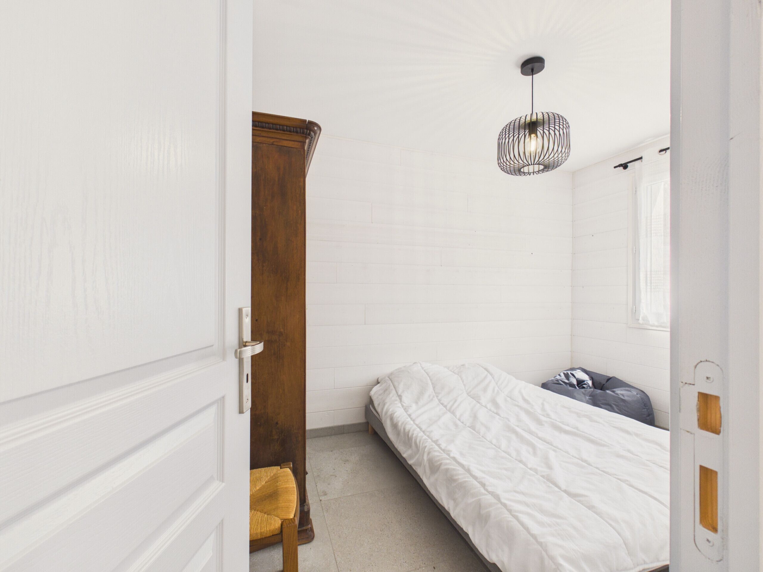 A bedroom with a bed, a wooden wardrobe, a chair, and a hanging light fixture.