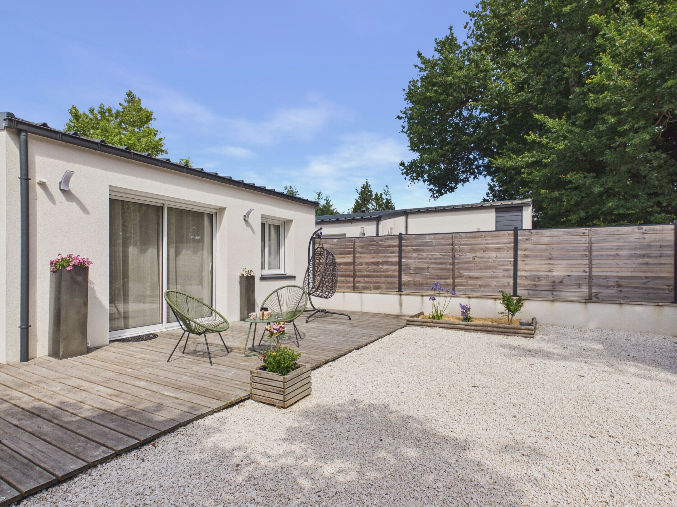 A modern outdoor patio with a wooden deck, two green chairs, a hanging chair, and a small garden area with gravel and plants.