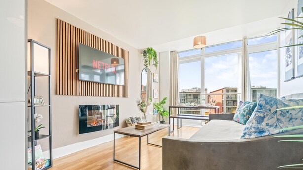 A modern living room with a flat-screen TV, wooden panel wall, large windows, and a balcony with a view of the city. - 1-bed Apartment, Sleeps 6 With Balcony View in West Midlands (Birmingham)