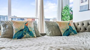 A bed with a textured white cover, two decorative pillows with a bird design, and a view of buildings through large windows. - 1-bed Apartment, Sleeps 6 With Balcony View in West Midlands (Birmingham)