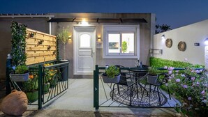 Ein modernes Haus mit geschlossenem Eingangsbereich, einer kleinen Terrasse mit Tisch und Stühlen sowie ein Garten mit Blumen und Pflanzen.