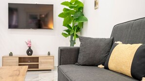 Ein modernes Wohnzimmer mit einem grauen Sofa, einem Holztisch, einem an der Wand montierten Fernseher und einer großen Grünpflanze.
