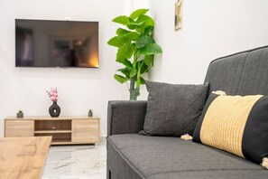 En moderne stue med en grå sofa, et træsidebord, et fjernsyn monteret på væggen og en stor, grøn plante.