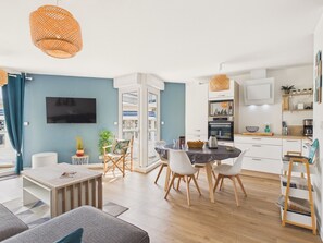 Ein modernes Wohnzimmer mit einer Couch, einem Couchtisch und einem Essbereich mit Stühlen. Es gibt eine Küche mit Schränken, einer Mikrowelle und einer Kaffeemaschine.