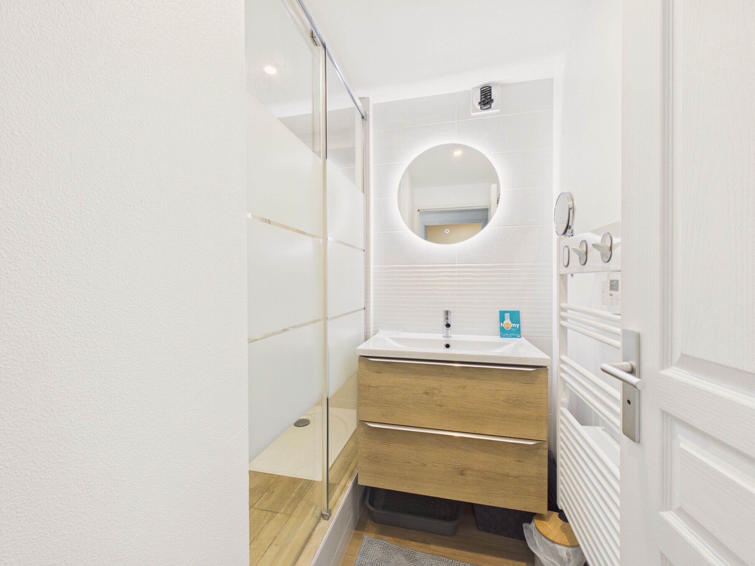 A modern bathroom with a glass shower enclosure, a round mirror, a white sink, and wooden cabinetry.