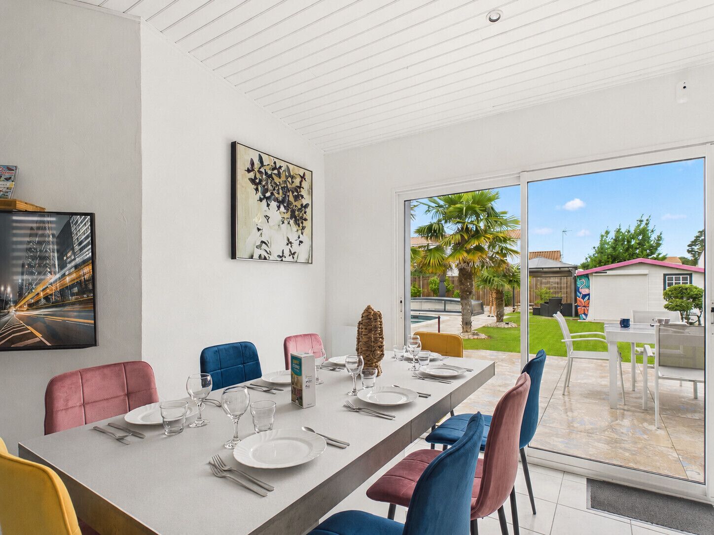 Uma sala de jantar com uma mesa comprida posta para quatro pessoas, rodeada por cadeiras, e uma grande janela com vista para um quintal com palmeiras.