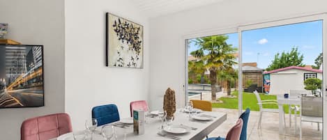 Un comedor con una mesa larga preparada para cuatro personas, rodeado de sillas y un gran ventanal que ofrece vistas a un patio trasero con palmeras.