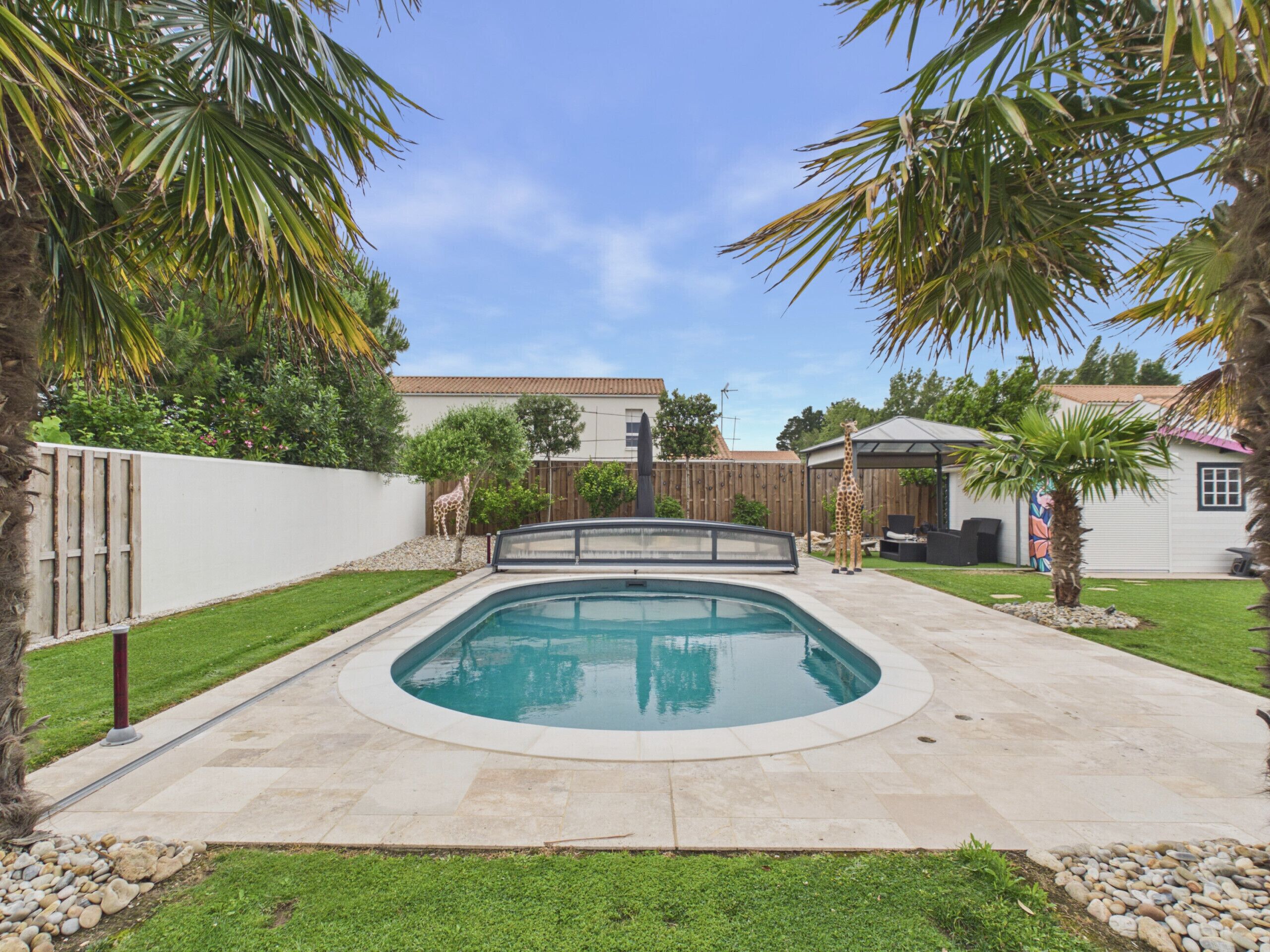 Uma piscina com deck de madeira, cercada por cerca branca, palmeiras e uma casa ao fundo.