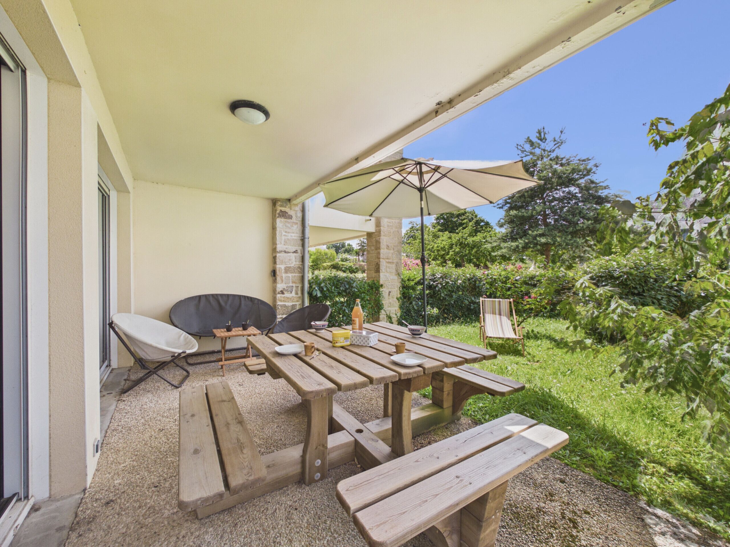 Une terrasse extérieure avec des tables et des bancs en bois, un barbecue et un grand parasol pour faire de l’ombre.