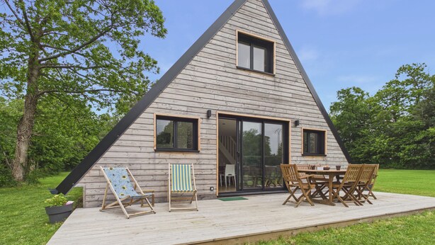 Una casa de madera triangular con terraza, mobiliario exterior y un amplio ventanal corredero.