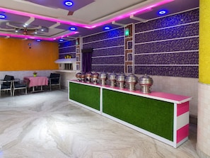 Interior moderno de restaurante com piso de mármore, balcão com recipientes de comida e iluminação azul e rosa no teto.